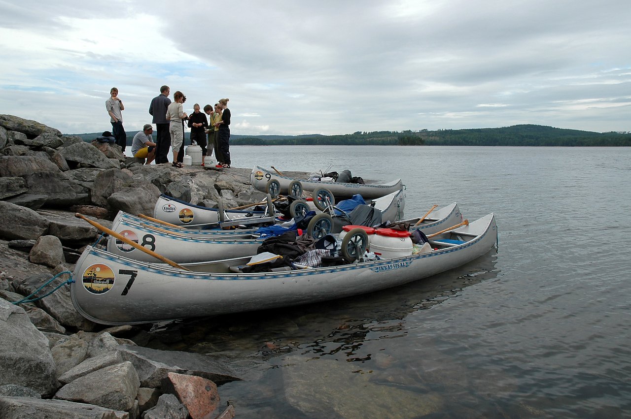 Several canoes loaded with gear are docked on a rocky shore while a group of people gathers nearby.