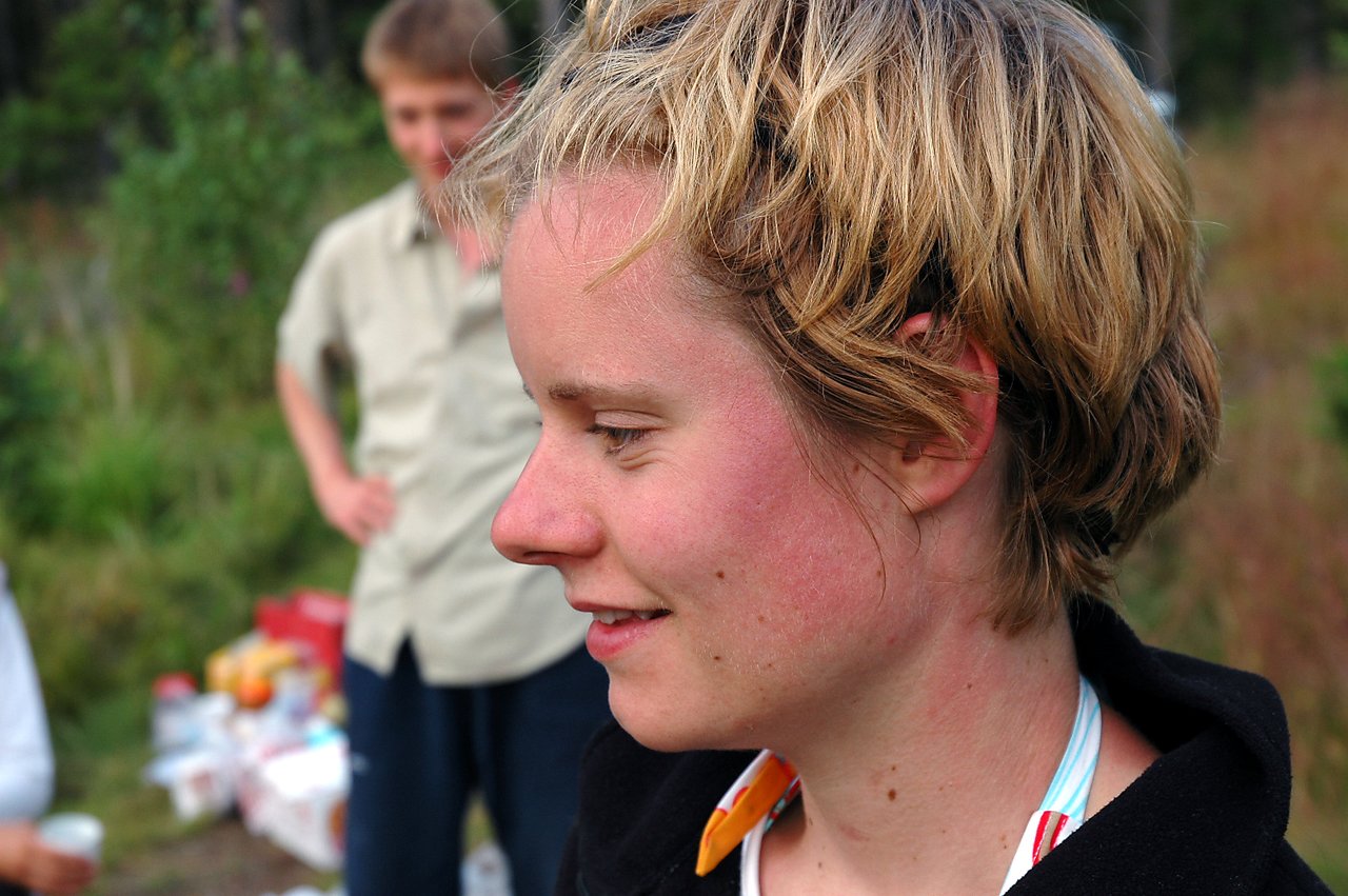 A woman smiles while looking to the side, with a man standing in the blurred background outdoors.