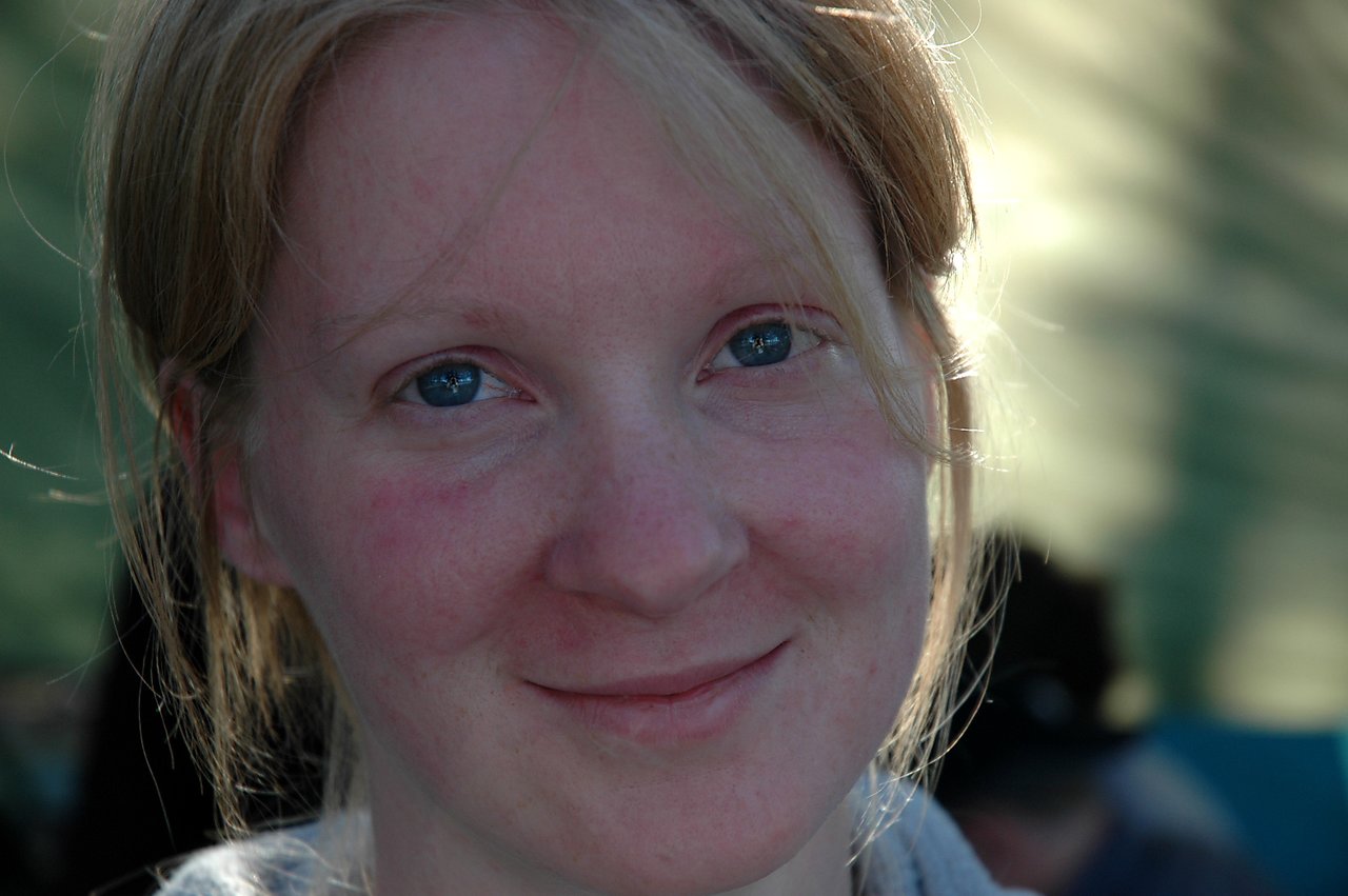 A woman with light skin and blue eyes smiles gently while looking at the camera in natural lighting.