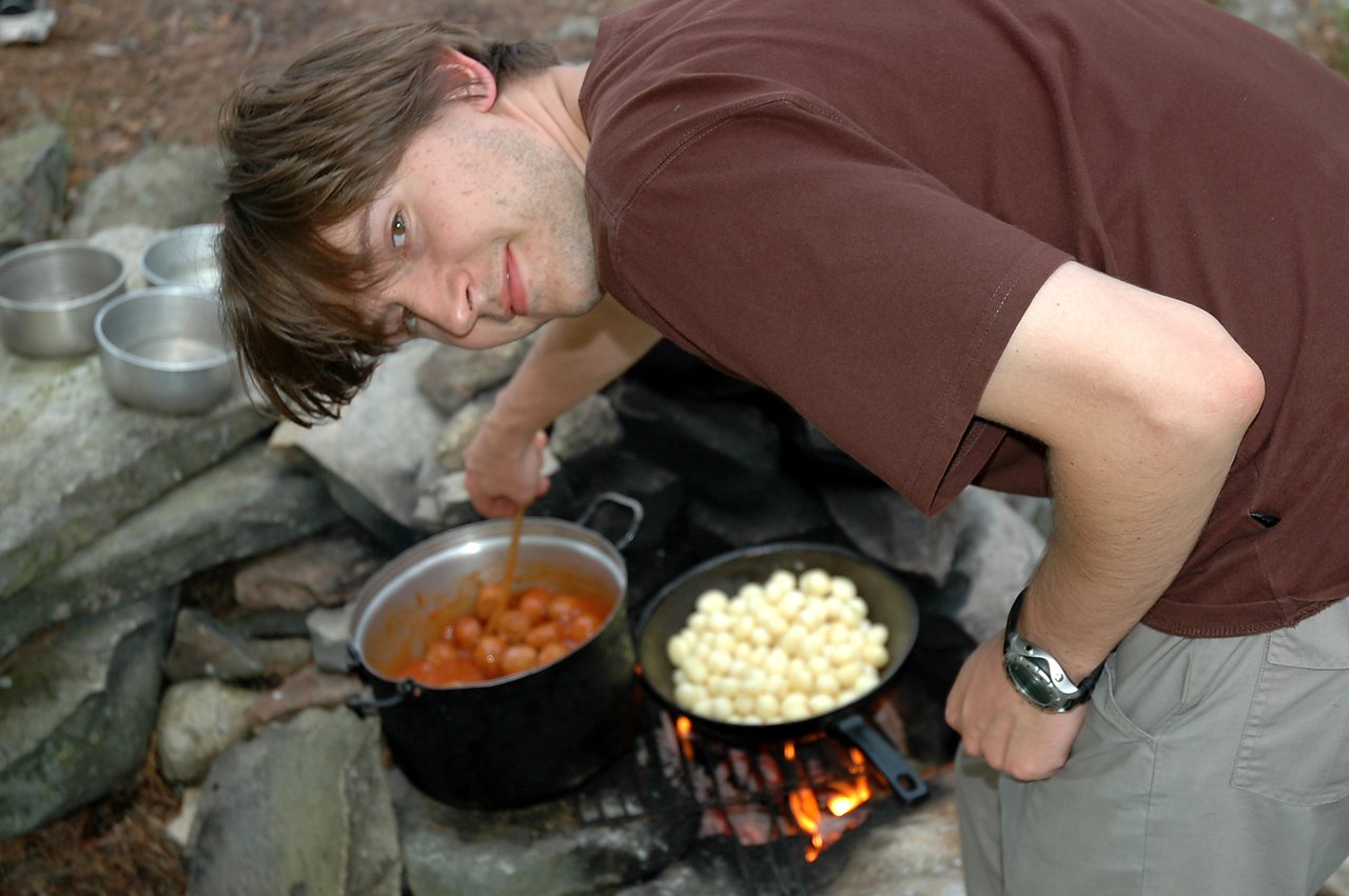 A man in a brown shirt stirs a pot of food over an outdoor fire, smiling at the camera.