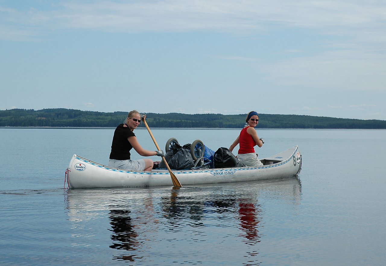 Two women paddle a canoe on a calm lake, carrying bags and a cart, both smiling at the camera.