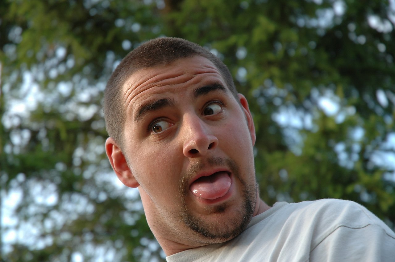A man in a light-colored shirt makes a playful face, sticking out his tongue and looking sideways.