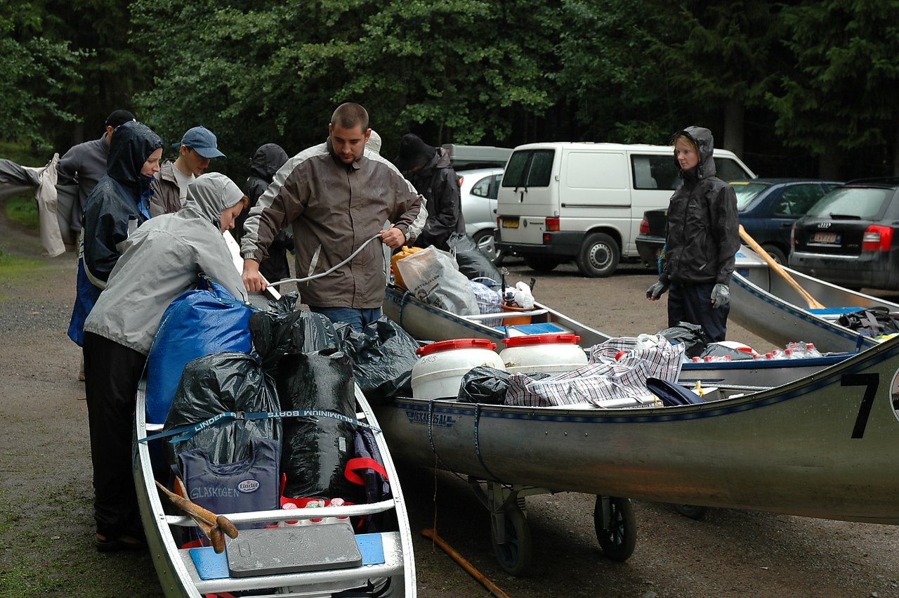 People in rain jackets pack canoes with bags, barrels, and supplies in a wet outdoor area.