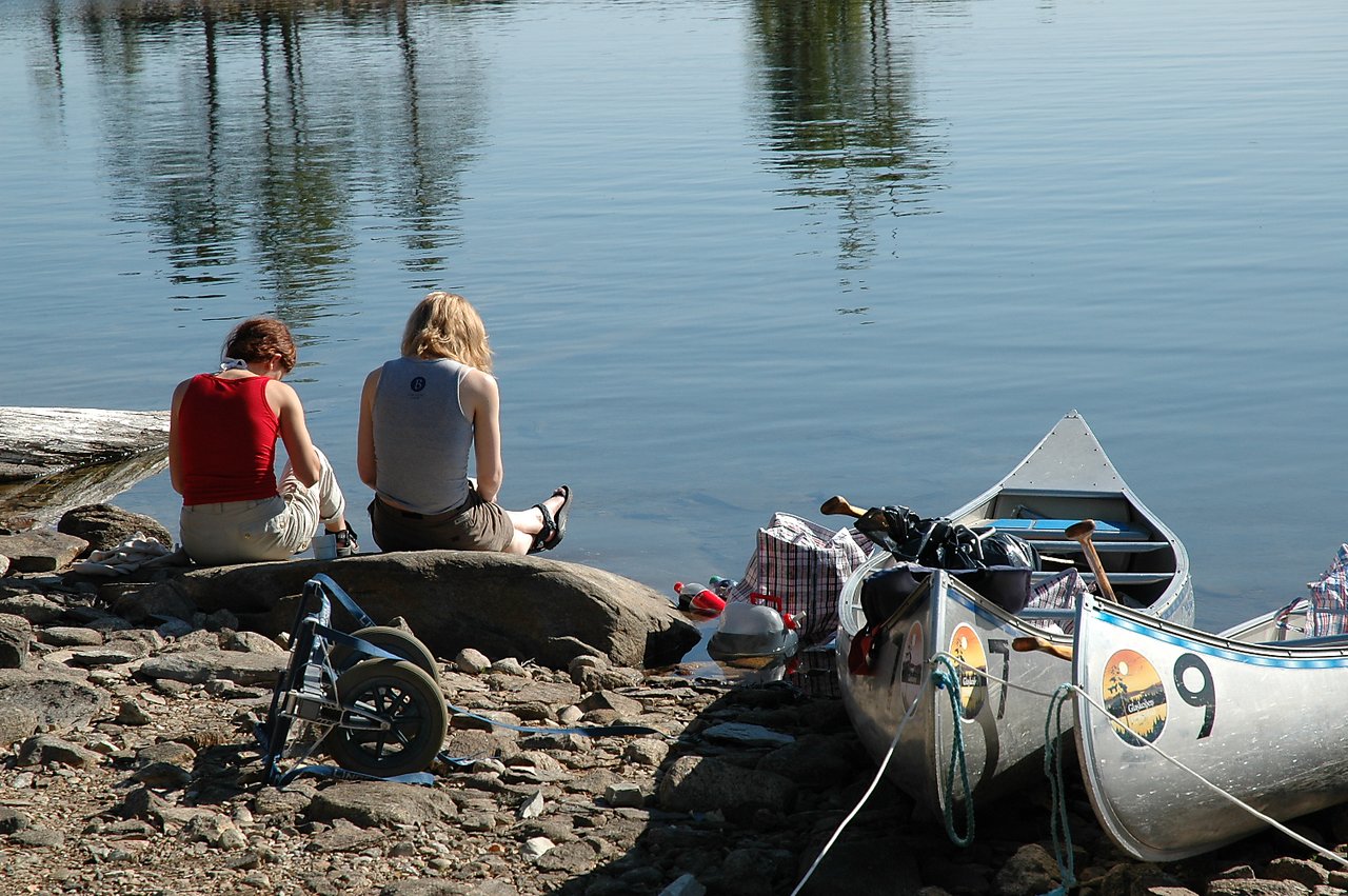 Two people sit on a rock by the water, reading a book together.