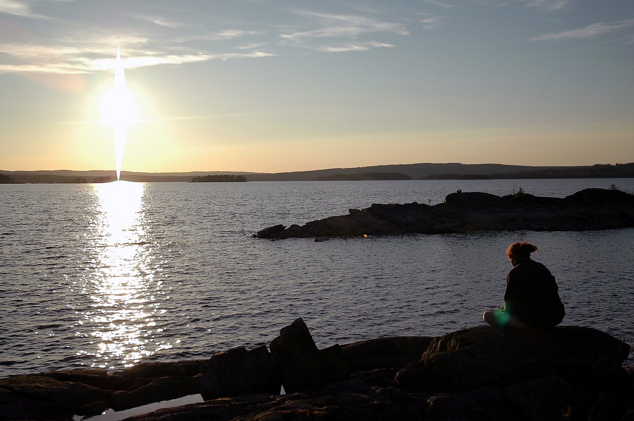 A person sits on a rocky shore, looking at the water during sunset.