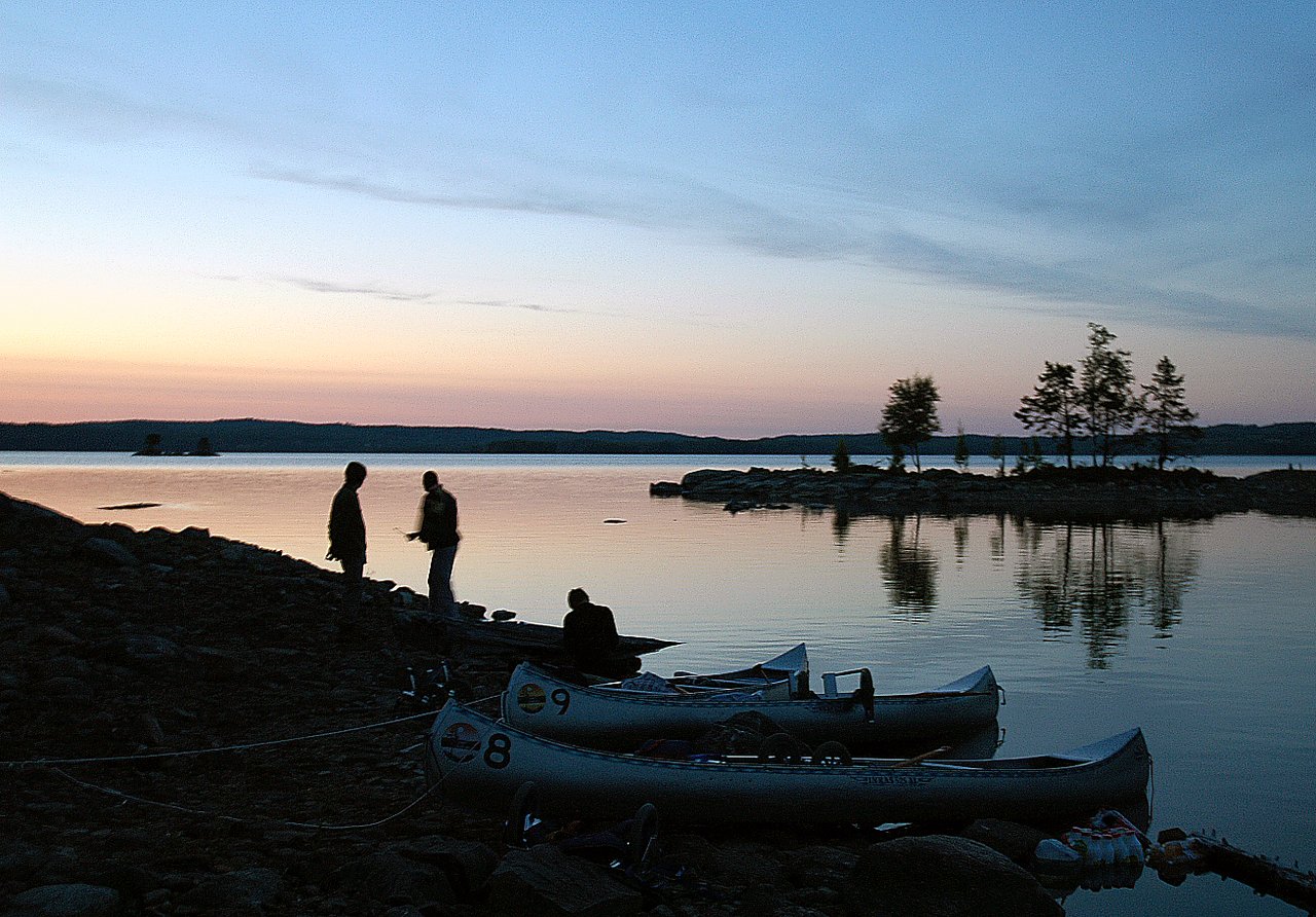 Two silhouetted people stand by the water, talking near canoes at dusk.