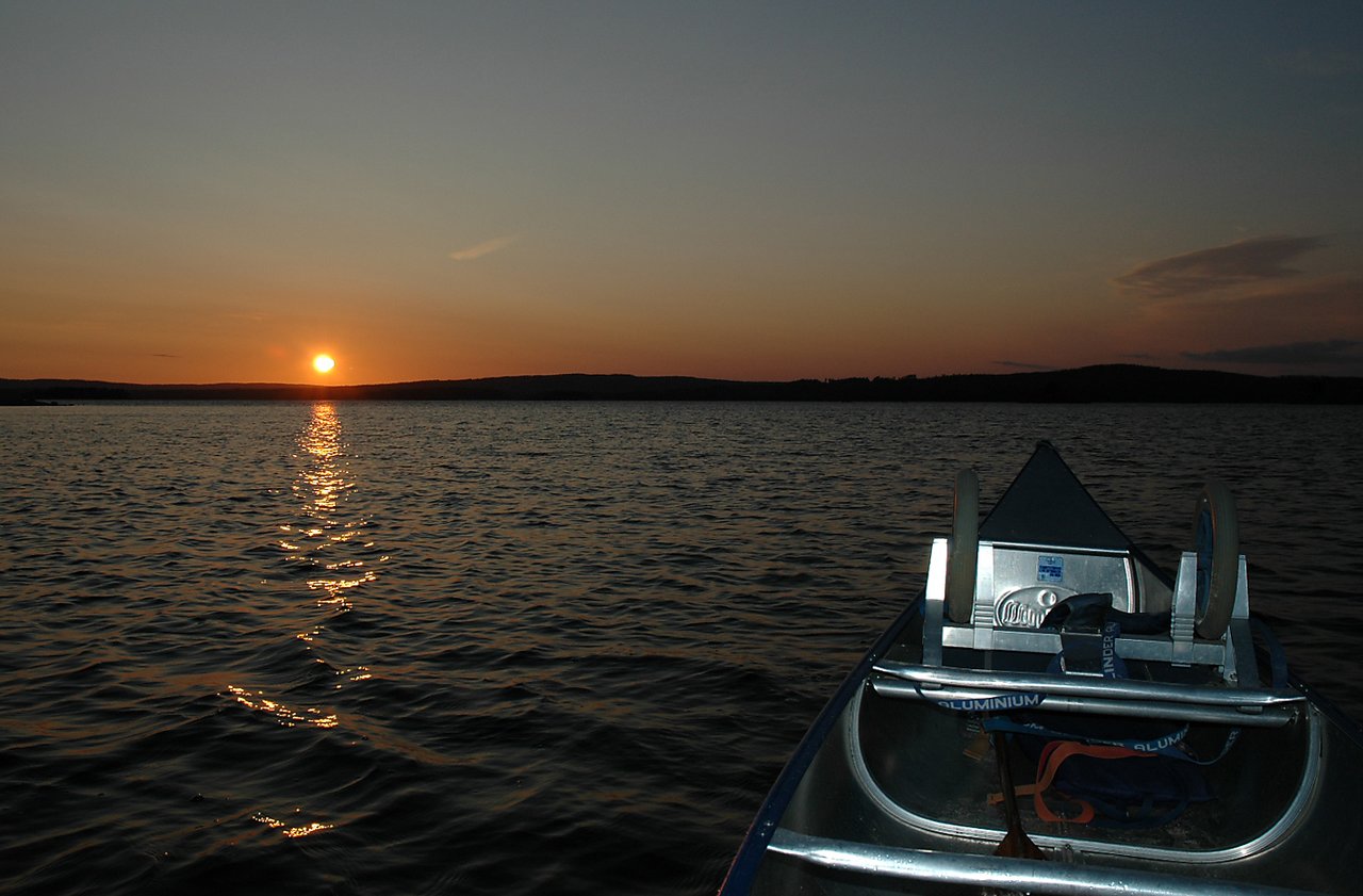 A canoe floats on a lake at sunset, with the sun reflecting on the water and distant hills in the background.