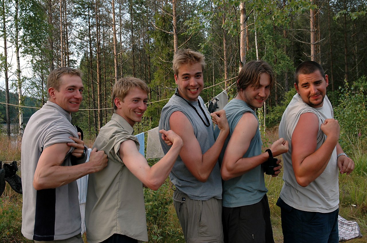 Five young men stand in a row outdoors, smiling and flexing their arms while pulling up their sleeves.