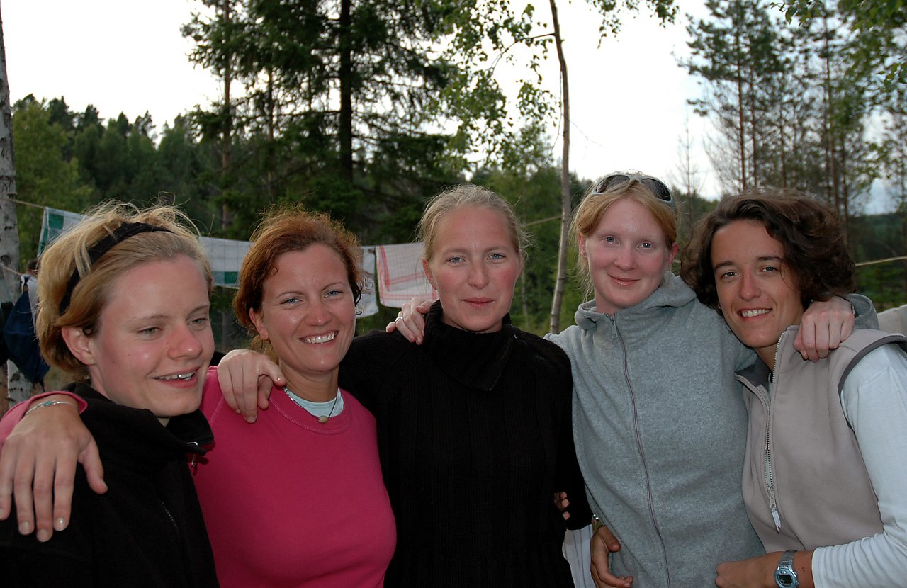 Five women stand close together outdoors, smiling and embracing each other with their arms around one another.