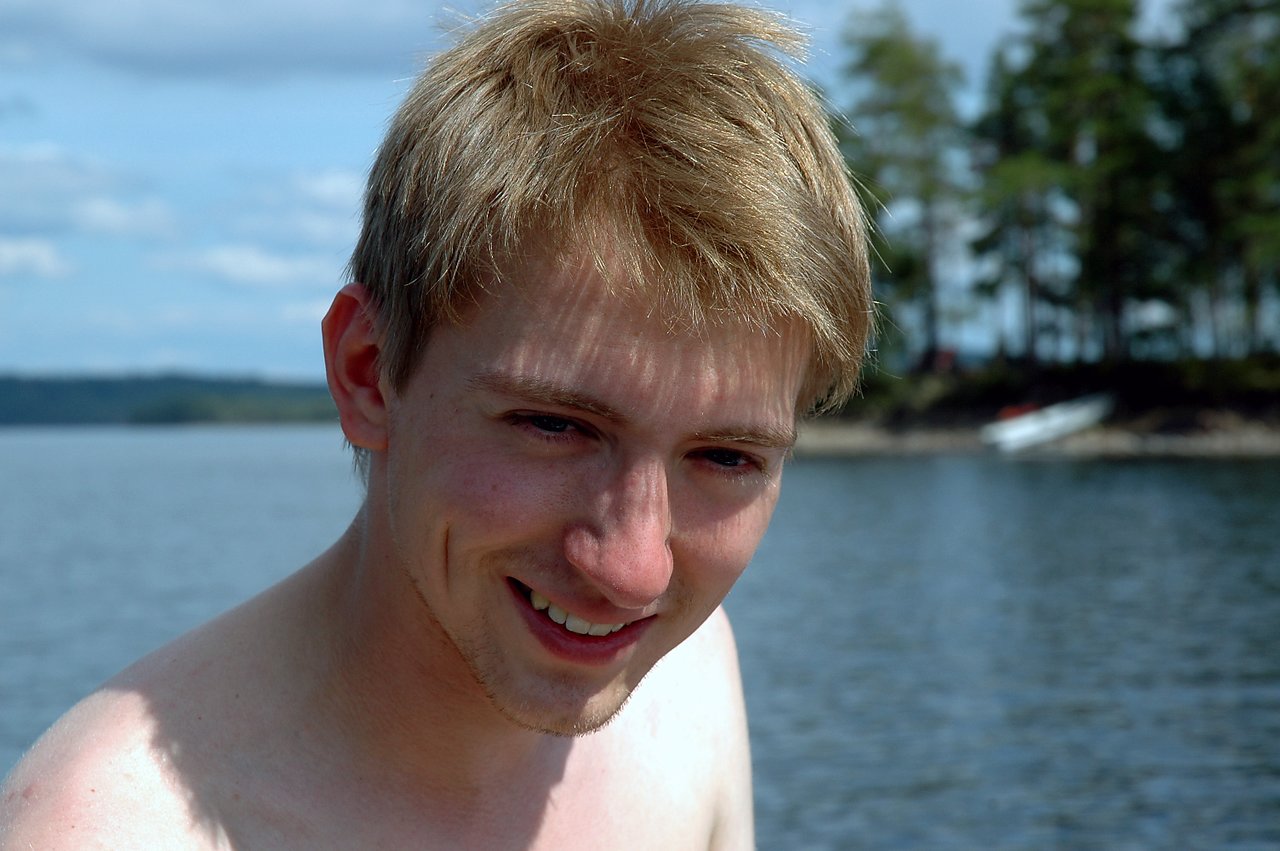A young man with short blond hair smiles while standing near a lake with trees in the background.