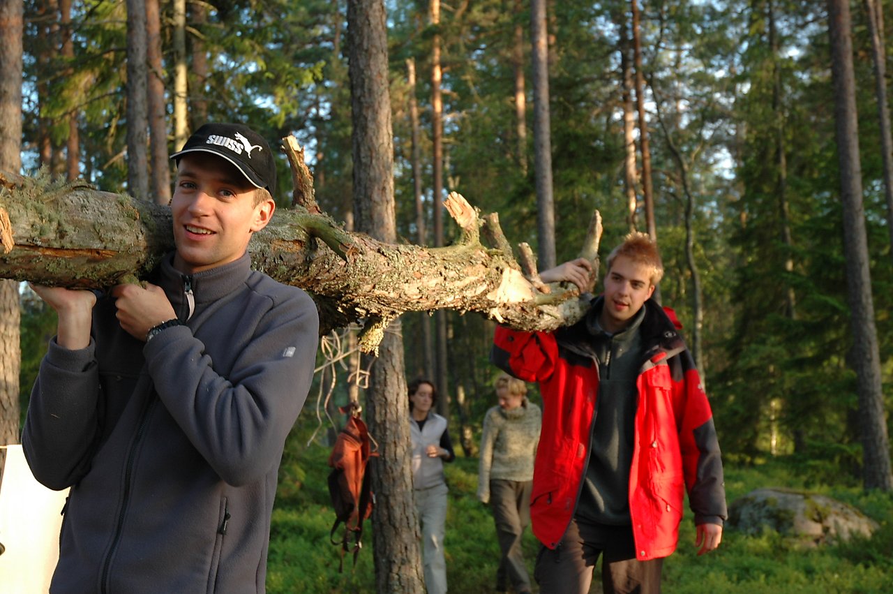 Two people carry a large tree branch through a forest, gathering wood for cooking.