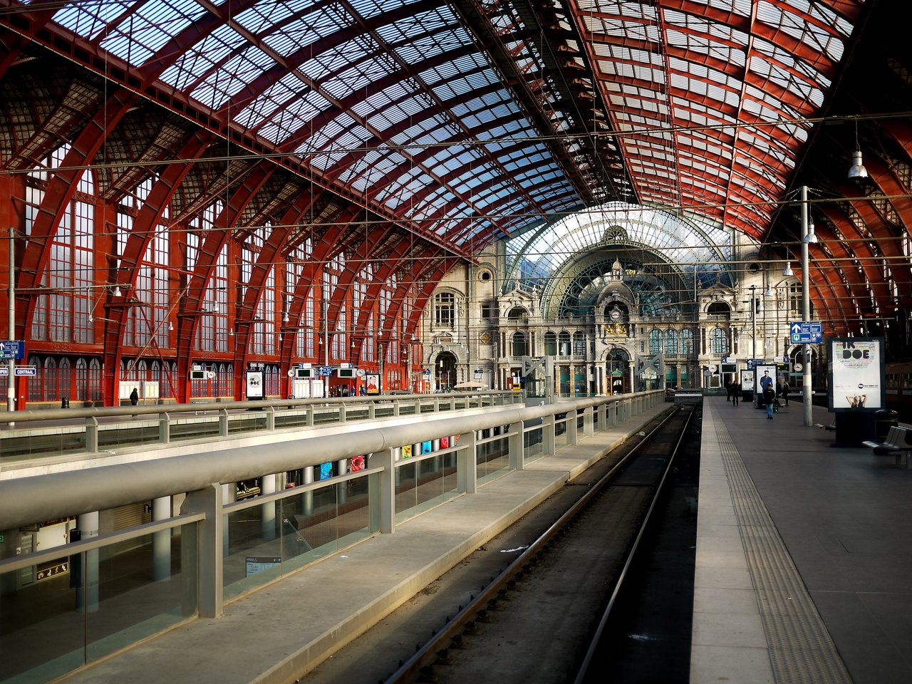 A spacious train station with a glass and metal roof, empty tracks, and a few people walking on the platform.