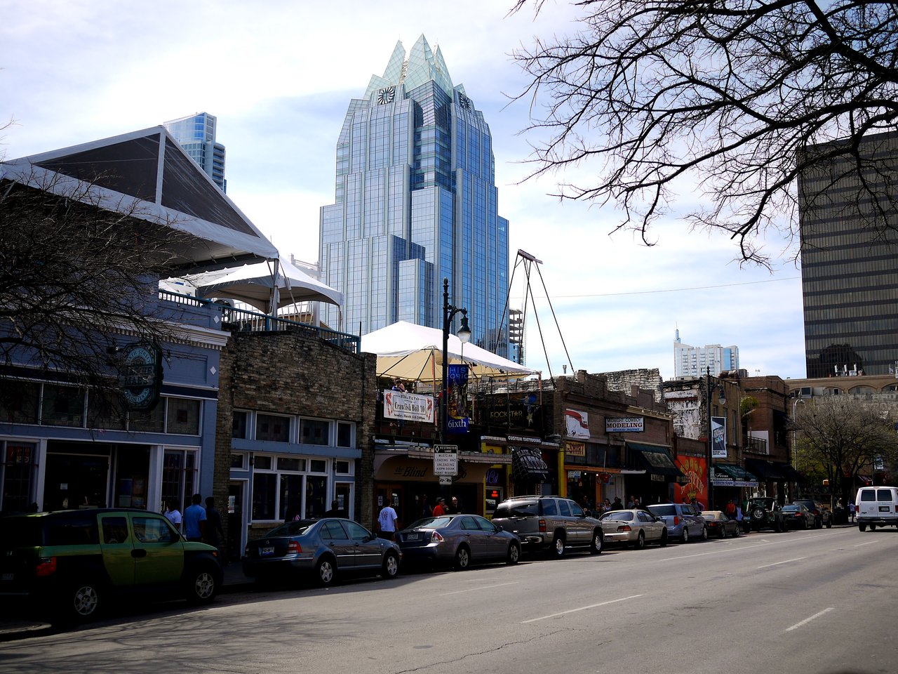A busy street in Austin with parked cars, people walking, and a tall glass building in the background.