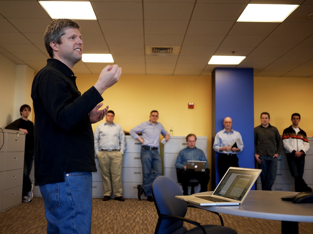 A man in a black shirt speaks to a group of people standing in an office setting.