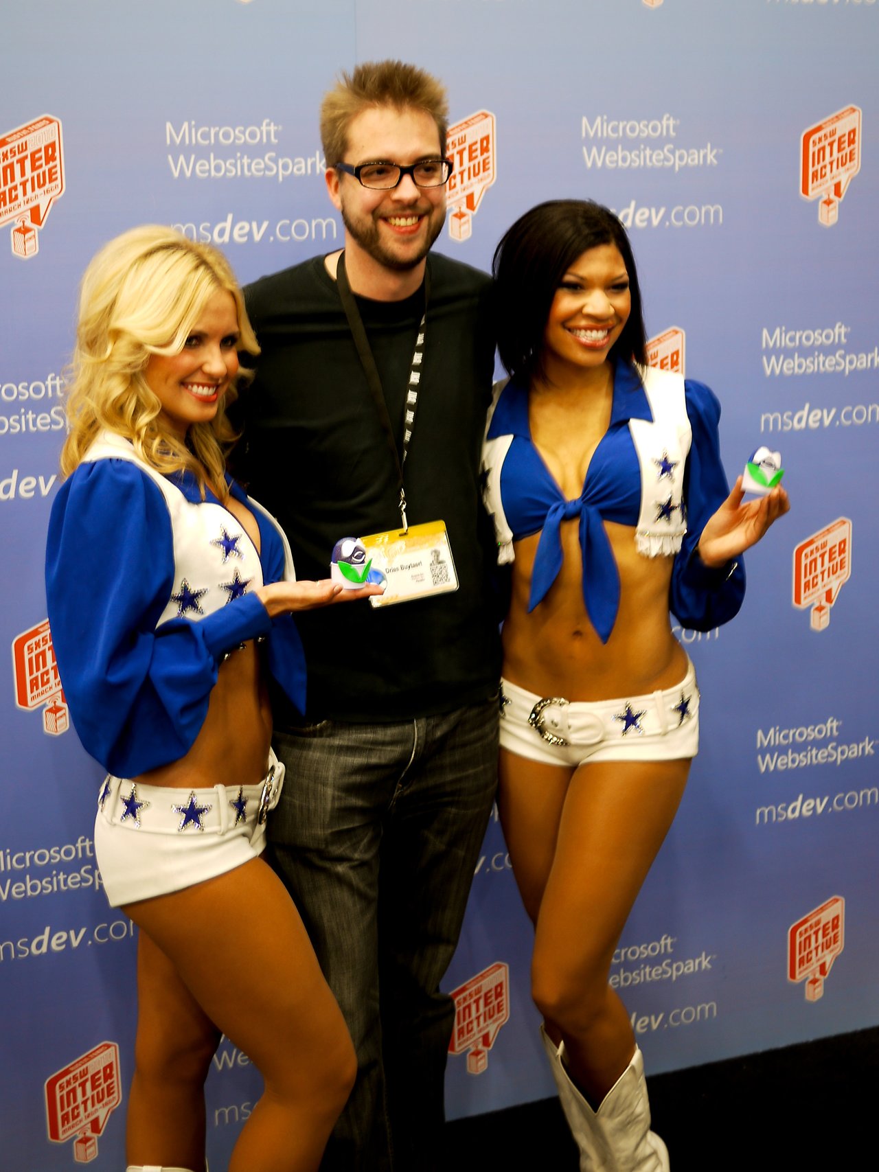 A man poses with two Dallas Cowboys cheerleaders in blue and white uniforms at an event, all smiling.