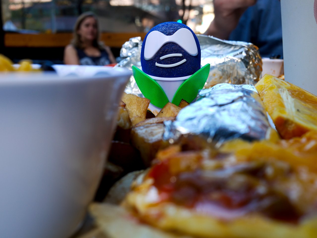 A small Drupal-themed toy stands among breakfast food, including foil-wrapped items, potatoes, and fruit, at an outdoor table.
