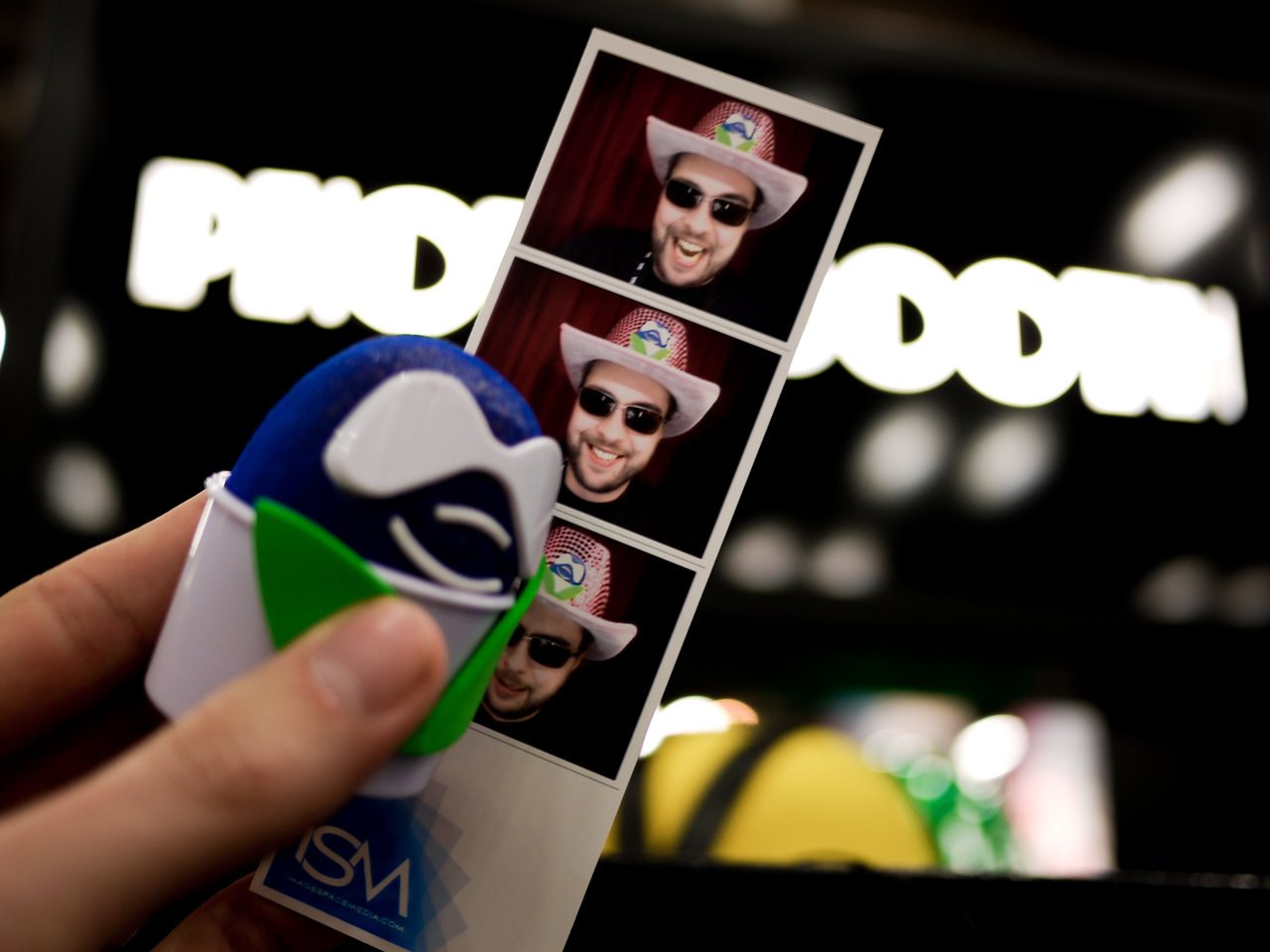 A hand holds a Drupal-themed toy and a photo strip of a smiling person wearing a cowboy hat and sunglasses.