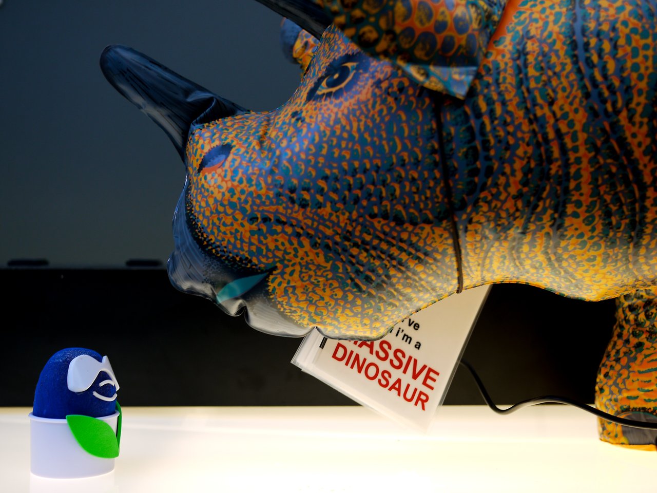 A large toy dinosaur holds a book in its mouth while facing a small blue and white figurine.