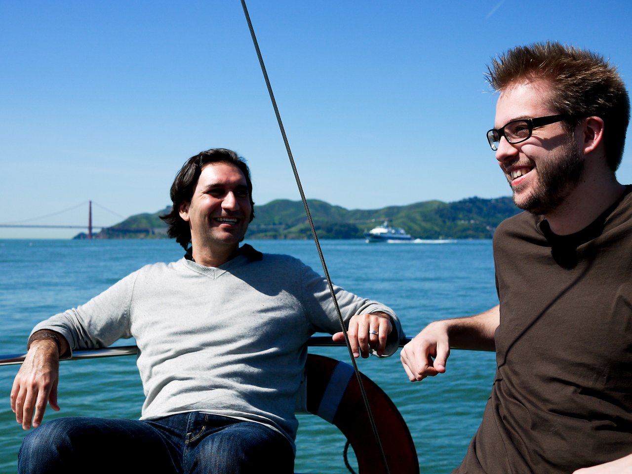 Two men sitting on a boat, smiling and talking, with water and a bridge in the background.