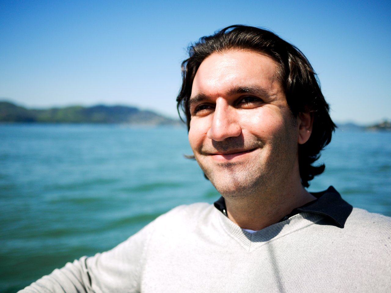 A person with dark hair and a light sweater smiles while standing outdoors near a body of water.
