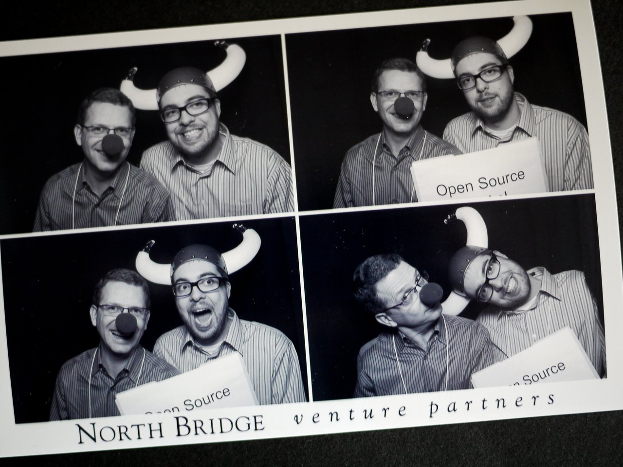 Two men in a photo booth wear playful props, including Viking hats and clown noses, holding a sign reading "Open Source.