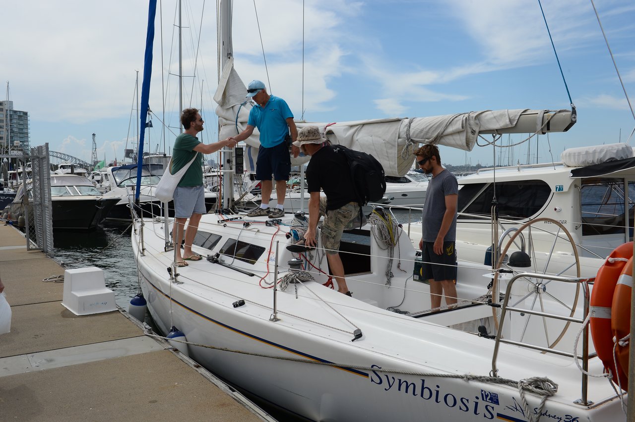Four men boarding a white sailboat docked at a marina; one is shaking hands of the captain.