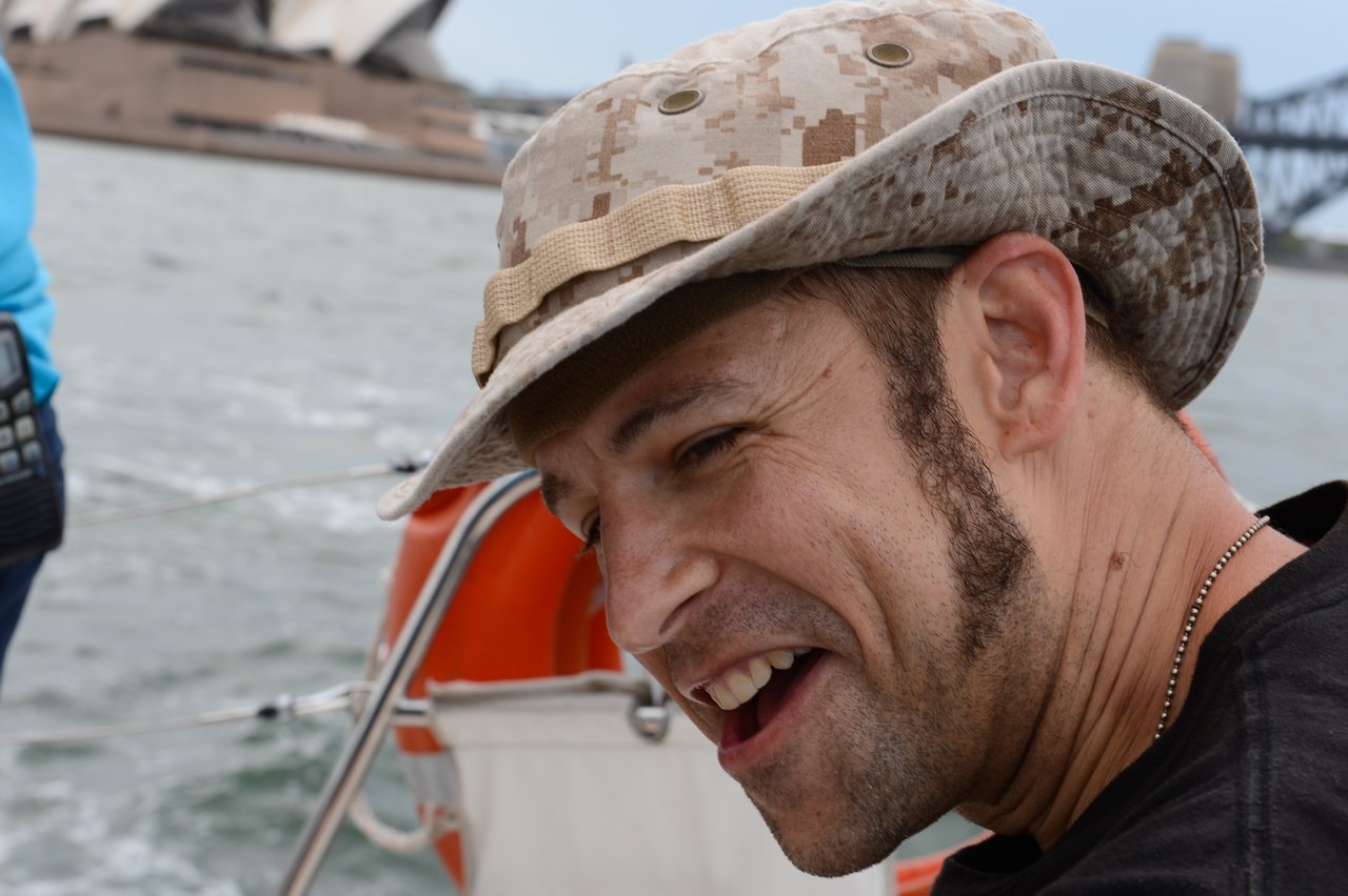 A man wearing a camouflage-patterned hat and black shirt smiles while leaning forward on a boat, with the Sydney Opera House in the background.