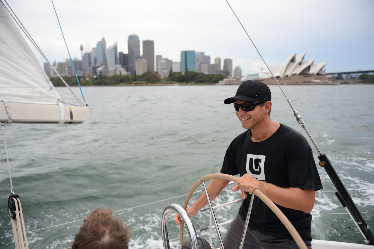A man wearing sunglasses and a black cap steers a sailboat with the Sydney Opera House and city skyline in the background.