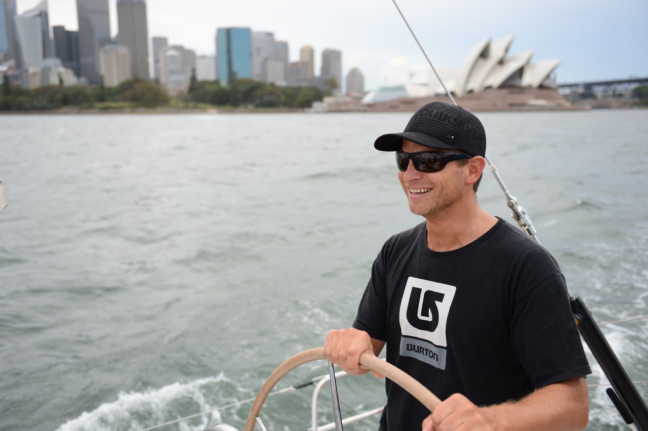 A man wearing a black cap, sunglasses, and a black t-shirt steers a sailboat on the water, with the Sydney Opera House and city skyline in the background.