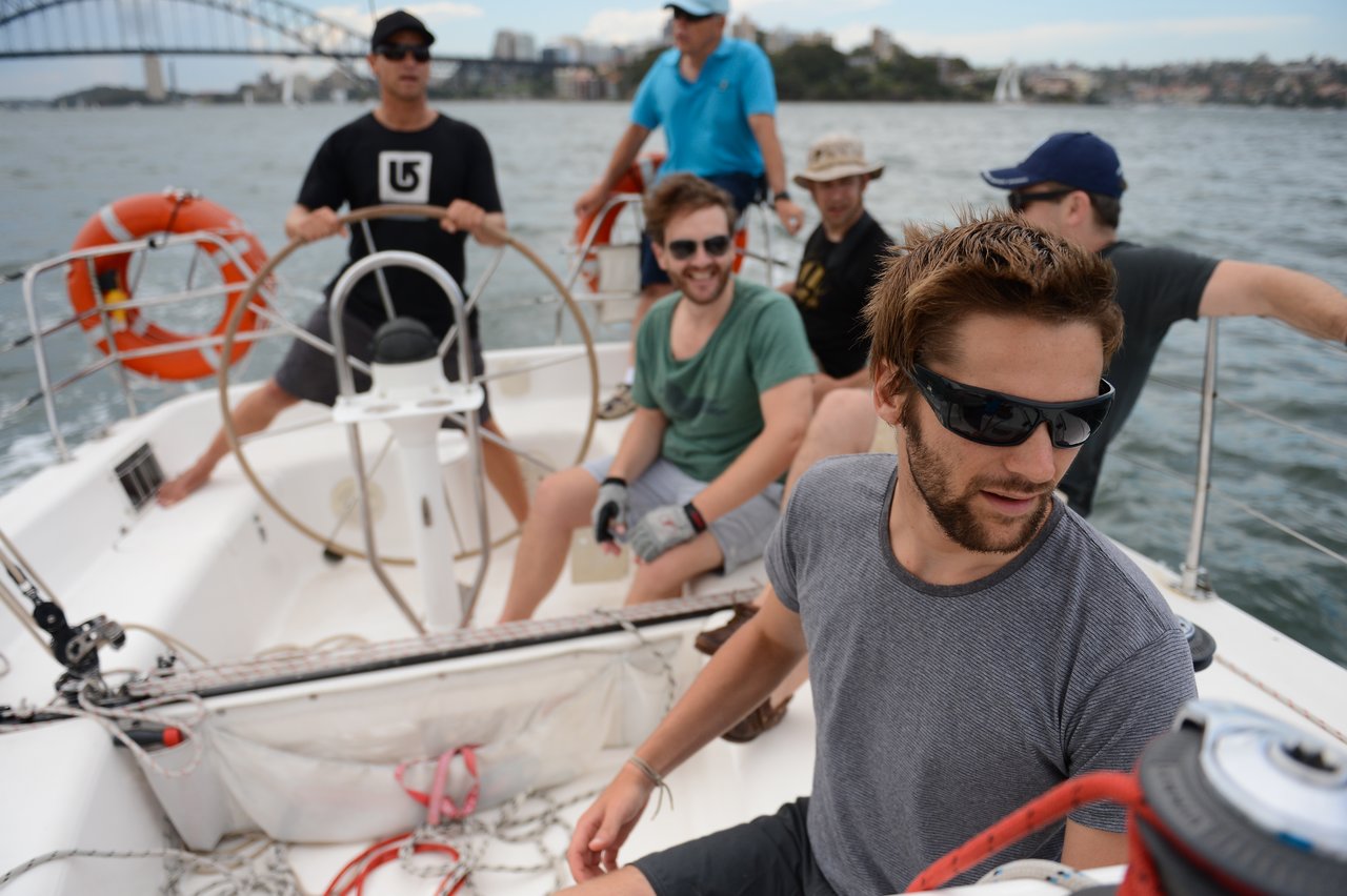 A group of six men are sailing on a boat, with one steering, others adjusting ropes, and the Sydney Harbour Bridge visible in the background.