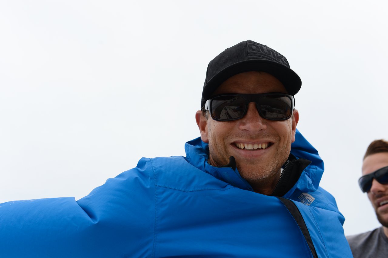 A man wearing a black cap, dark sunglasses, and a bright blue jacket smiles at the camera.