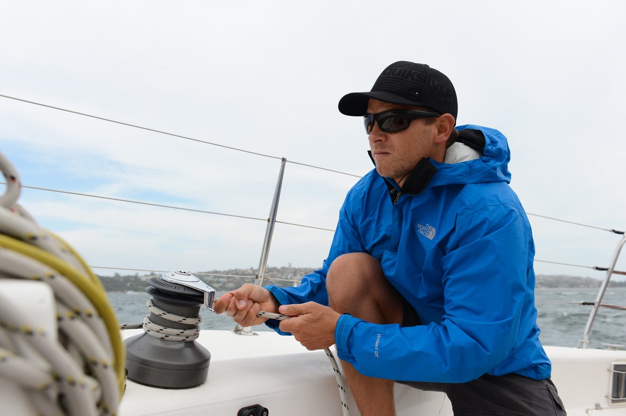 A man wearing a blue waterproof jacket, black cap, and sunglasses is crouched on a sailboat, gripping a rope and adjusting a winch.