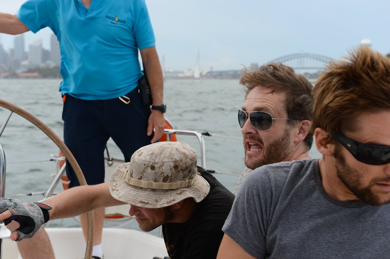Four men are on a sailboat, with one steering, another adjusting ropes, and two reacting intensely, while the Sydney Harbour Bridge and city skyline are visible in the background.