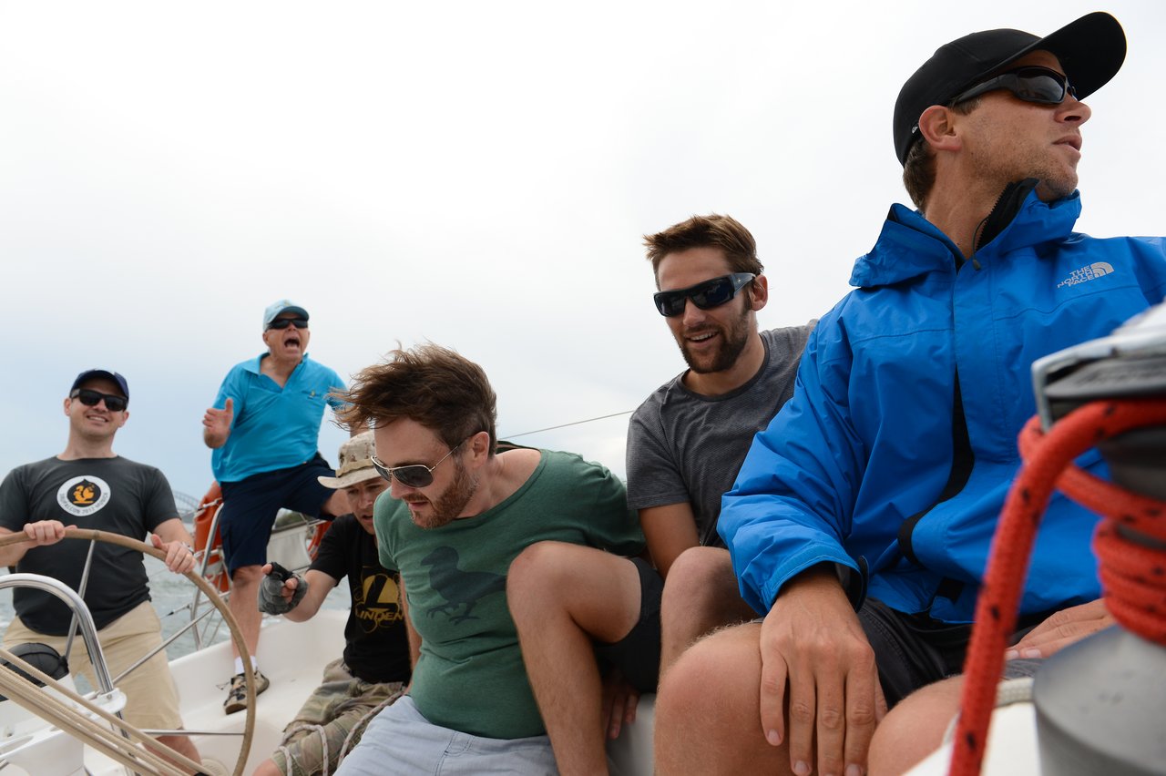 A group of six men wearing casual outdoor clothing and sunglasses are actively sailing a boat, leaning and adjusting positions as one steers.