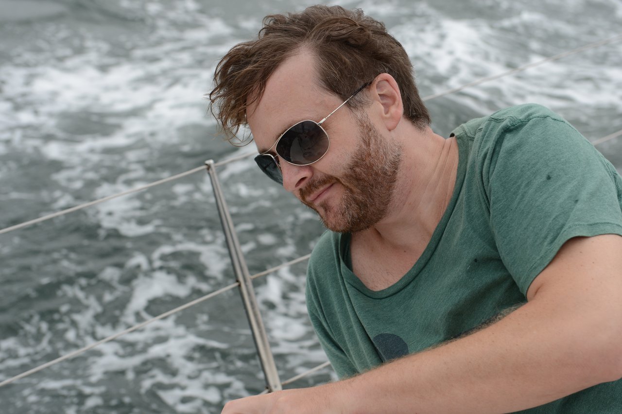 A man with short, wavy brown hair, a beard, and sunglasses, with ocean waves and a metal railing behind him.