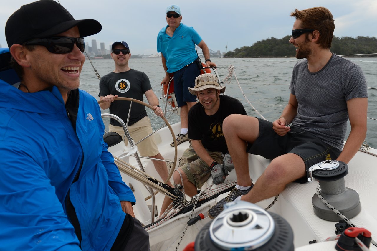 Five men are on a sailboat, smiling and laughing, with one steering and others adjusting ropes.