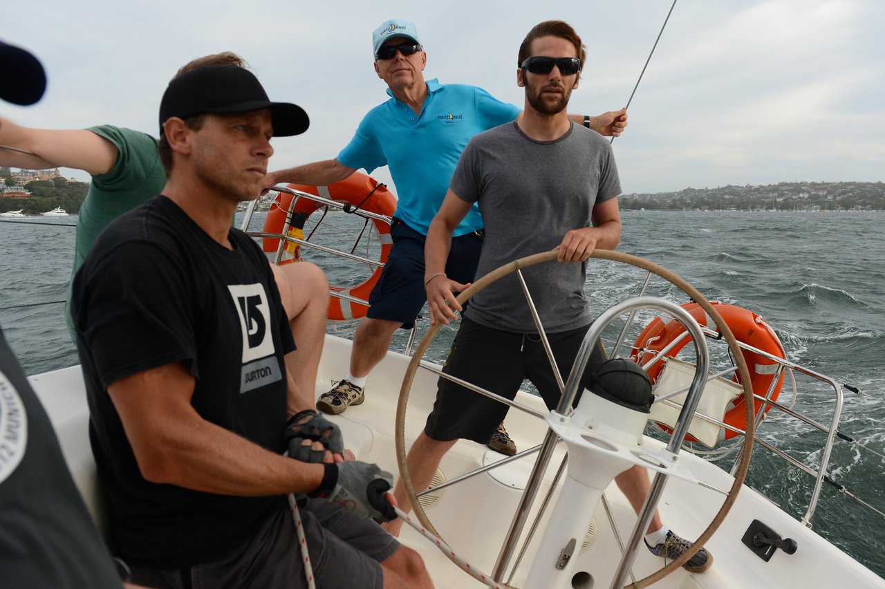 Three men are actively sailing a boat on choppy waters, with one steering the wheel, another handling ropes, and a third balancing near a lifebuoy.