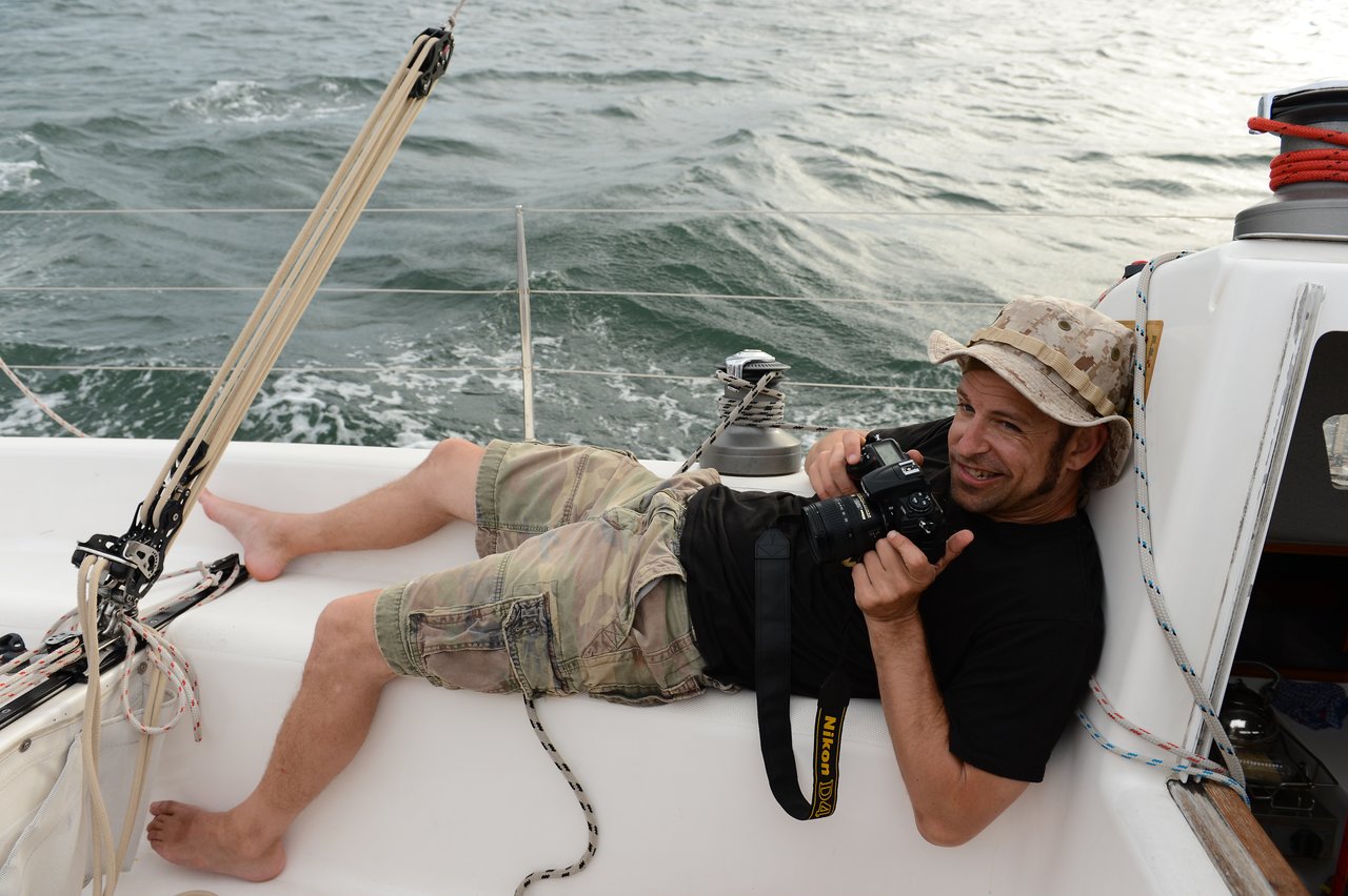 A man wearing a camouflage hat, black shirt, and cargo shorts reclines on a sailboat, holding a camera.