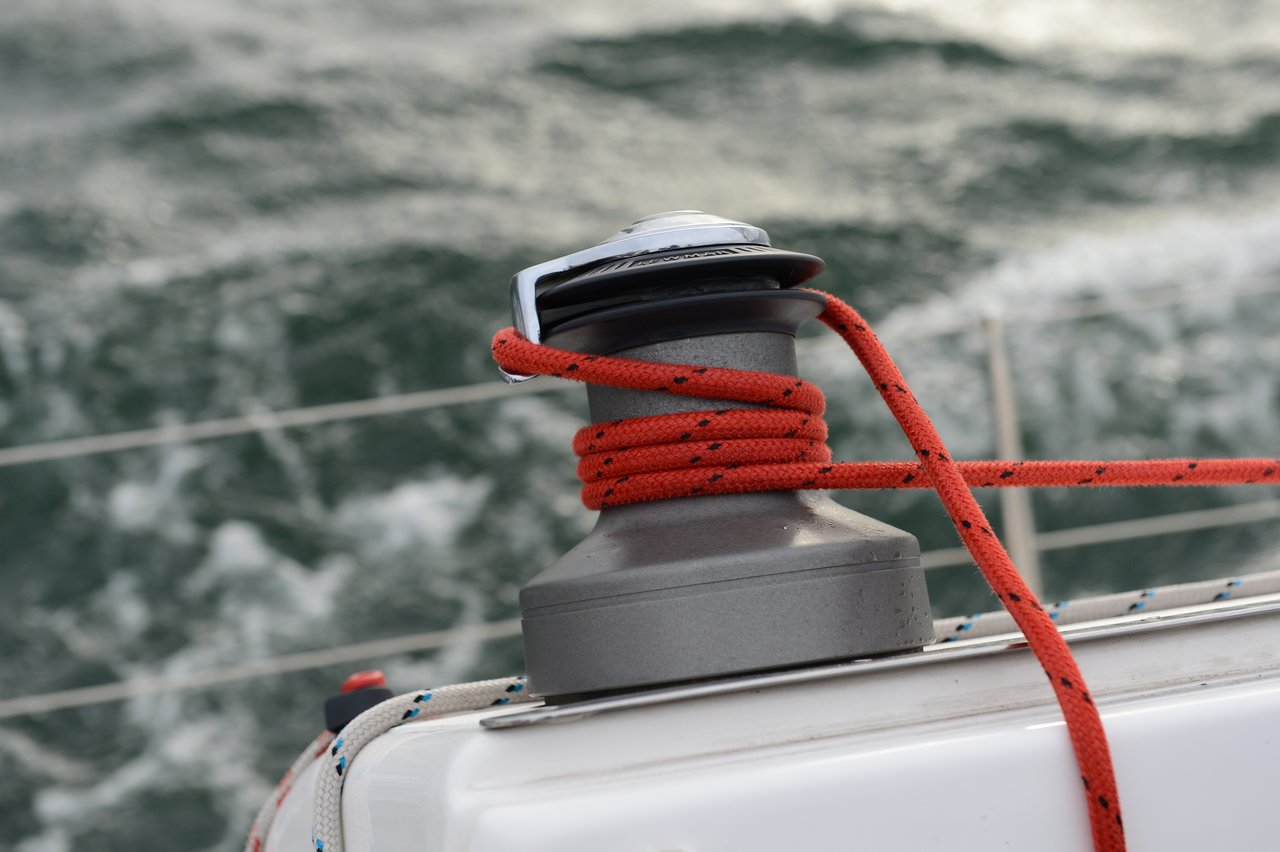 A close-up of a sailboat winch with a red rope tightly wrapped around it, set against a blurred background of choppy ocean waves.
