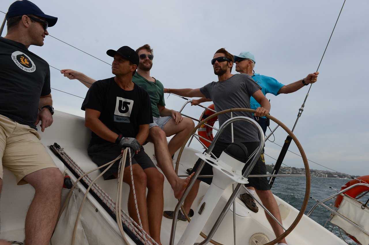 Five men on a sailboat, actively steering and handling ropes, with a tilted deck.