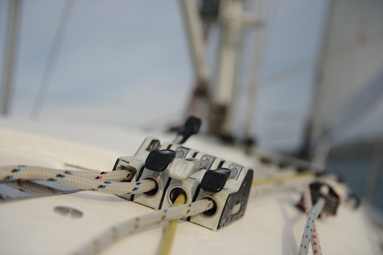 Close-up of a rope clutch on a sailboat deck, with multiple colorful ropes running through it, and a mast in the background.