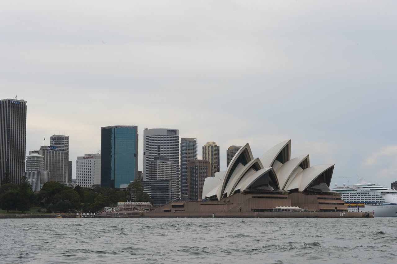 The Sydney Opera House with its distinctive white sail-like roofs near the waterfront, surrounded by modern high-rise buildings and a large cruise ship docked nearby.