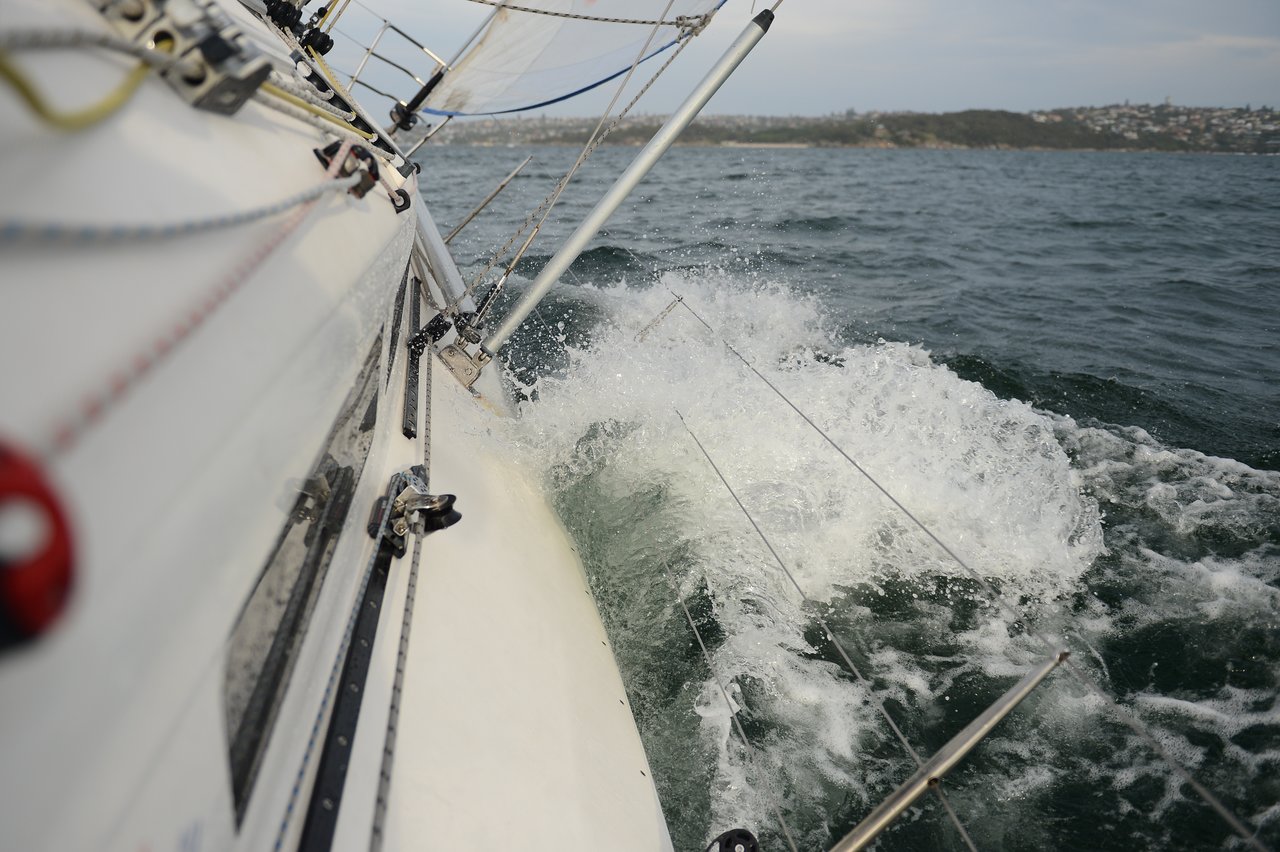 A close-up of a sailboat tipping sharply in rough water, with waves washing over the submerged railings.