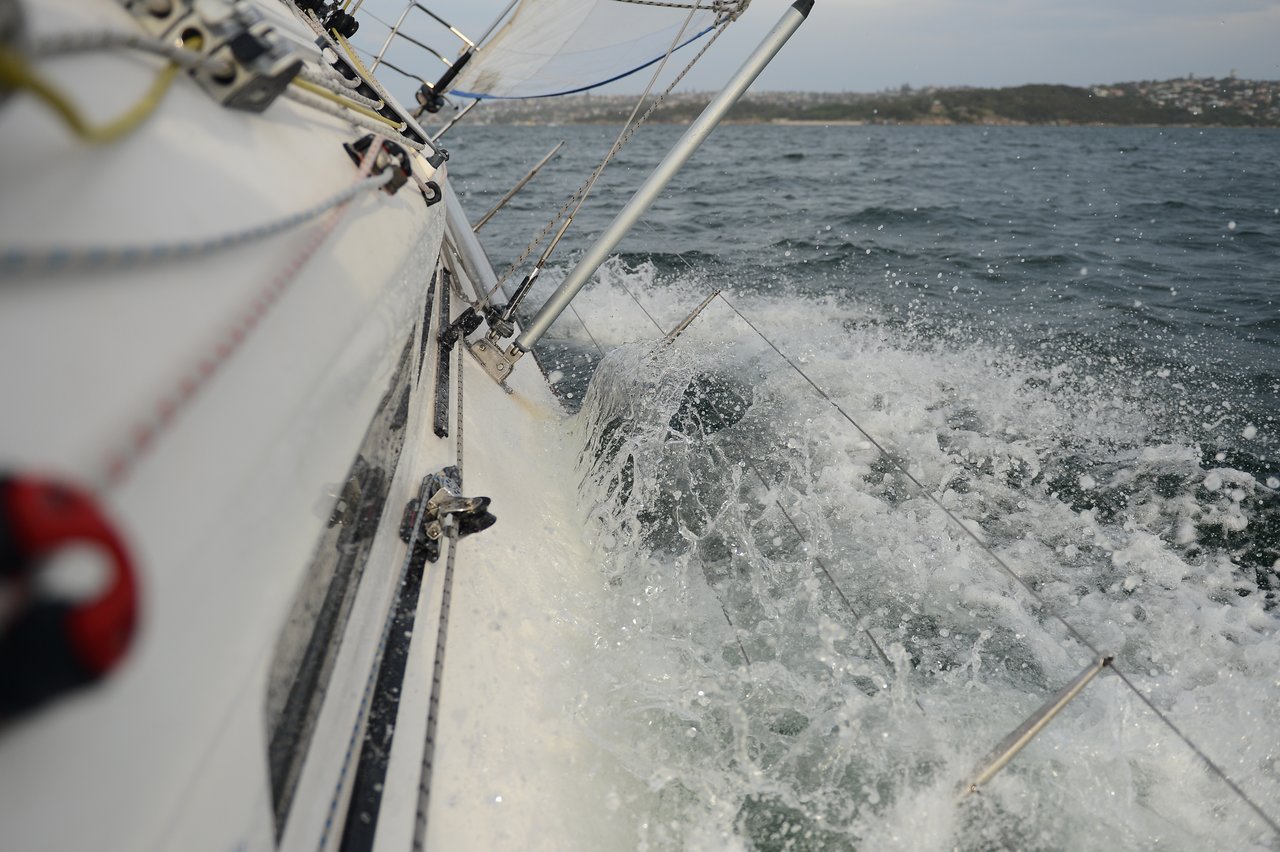 A close-up of a sailboat tipping sharply in rough water, with waves washing over the submerged railings.