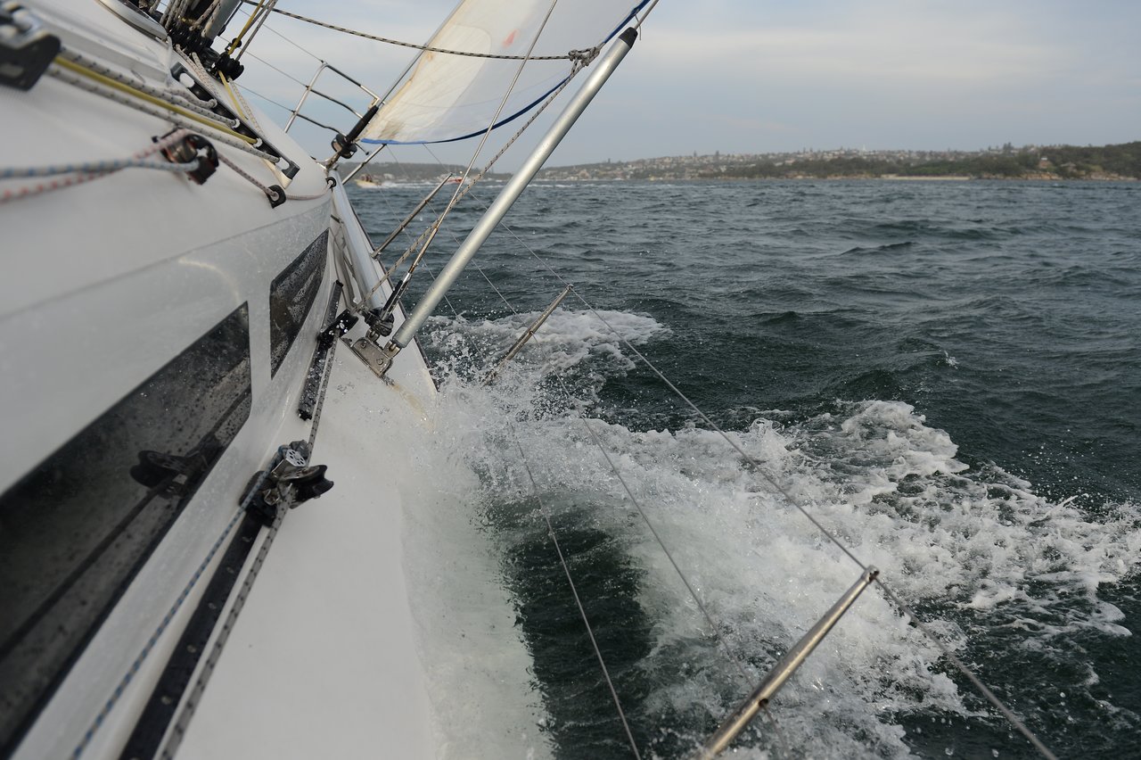 A close-up of a sailboat tipping sharply in rough water, with waves washing over the submerged railings.