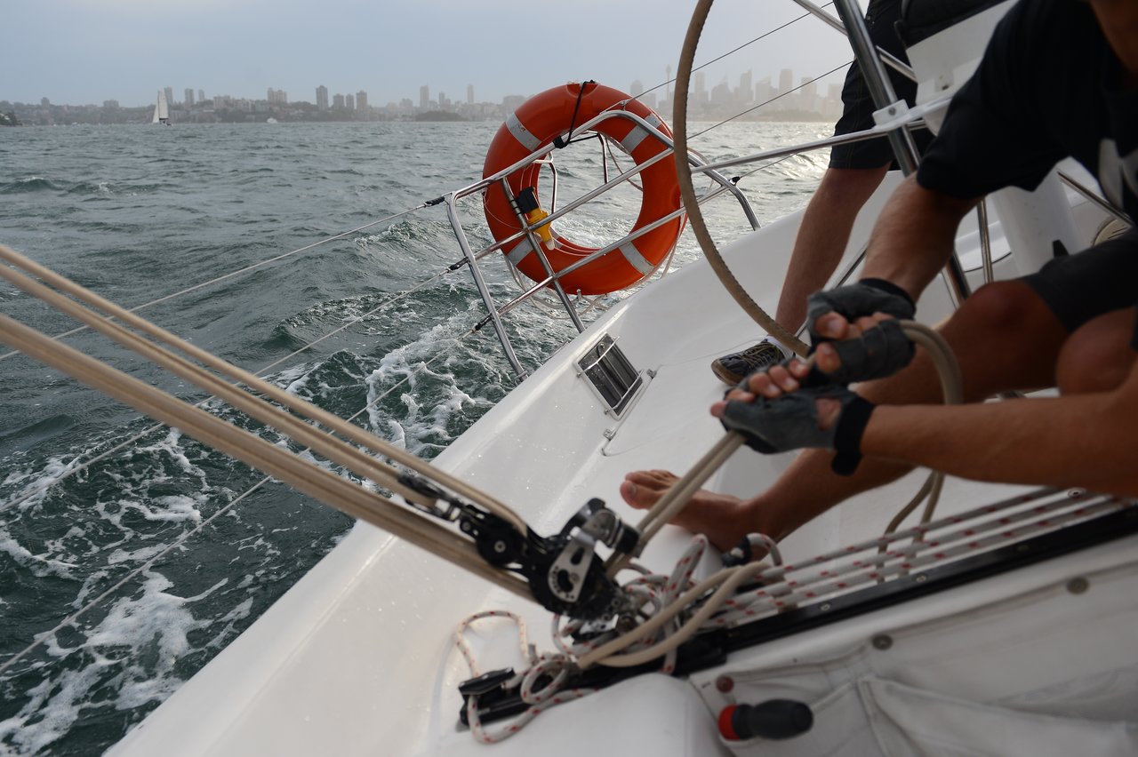 A person on a sailboat, wearing fingerless gloves, adjusts the ropes and pulleys while choppy water and a city skyline stretch out in the background.
