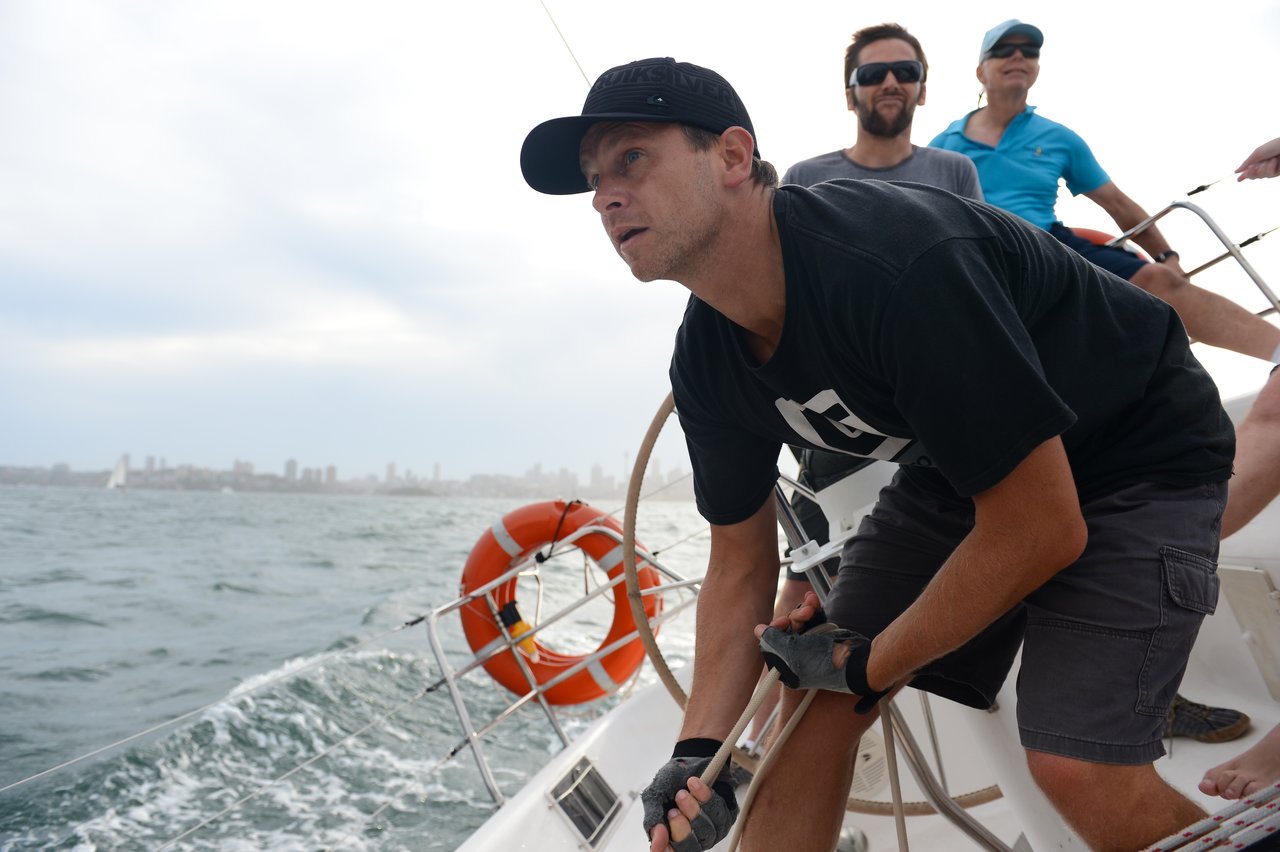 A man in a black cap and gloves grips a rope on a sailboat, leaning forward with focus as he navigates the choppy water, with a city skyline in the background.