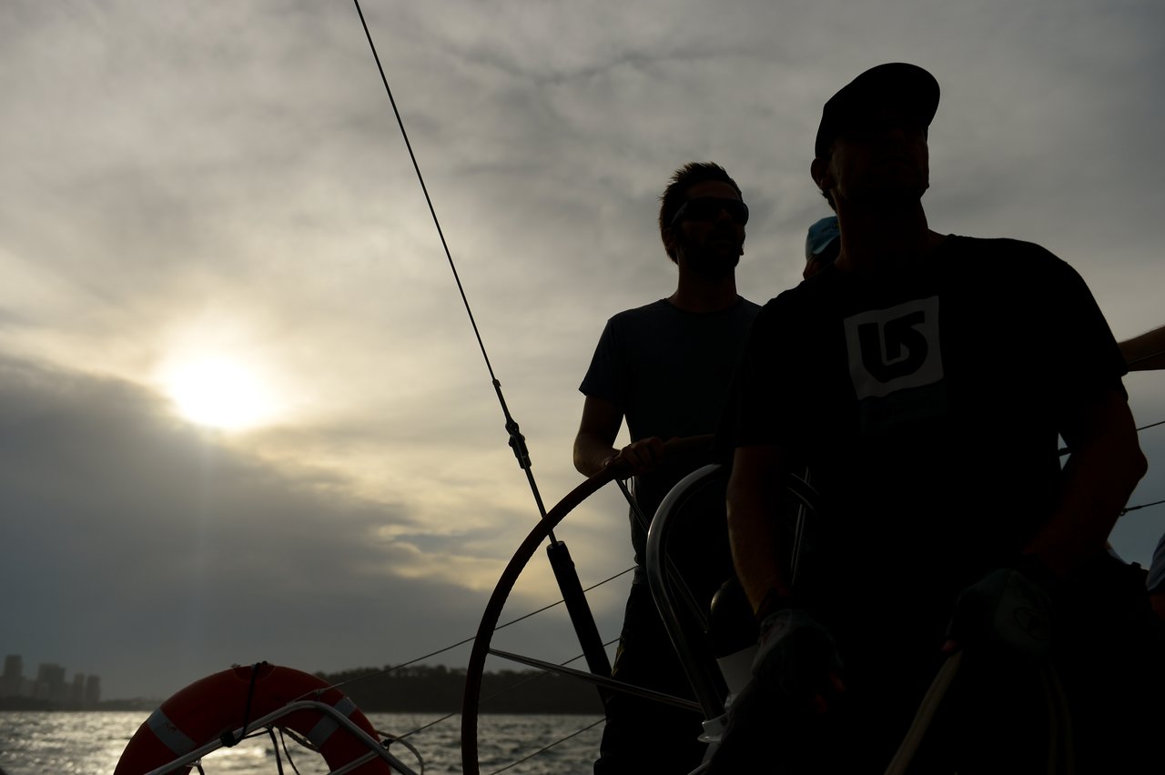 Silhouetted against a cloudy sky, two people are on a sailboat, with one steering the wheel, while the sun sets over the water.
