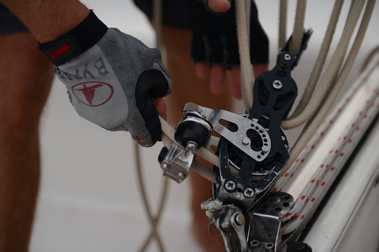 A person wearing fingerless sailing gloves grips a rope near a metal pulley system on a sailboat, with additional ropes and rigging visible in the background.