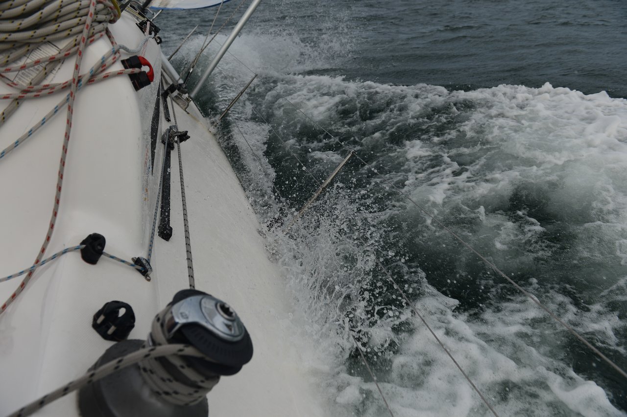 A close-up view of a sailboat's deck shows ropes, winches, and rigging as the boat tilts, cutting through choppy water with white foam splashing against the hull.